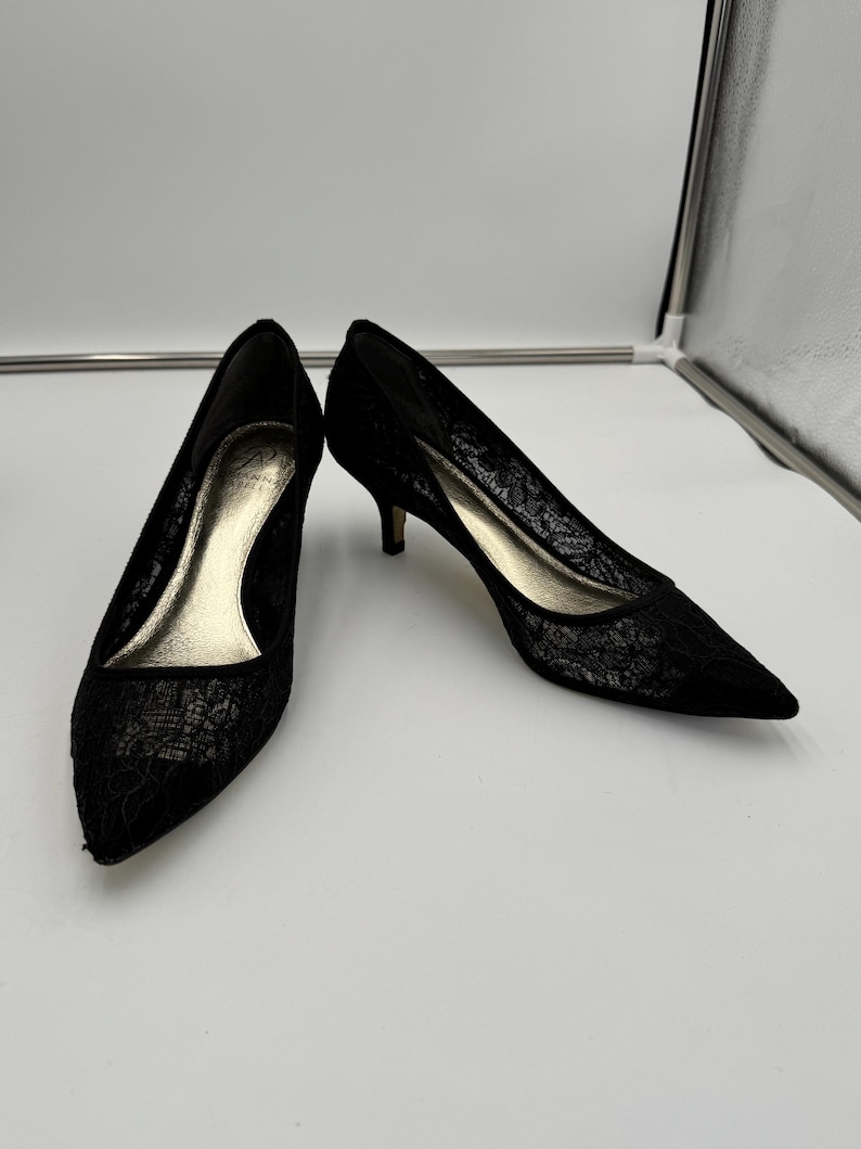 K&ouml;nnte beinhalten: Ein Paar schwarze Pumps mit Spitze, spitzer Zehenpartie und kleinen Abs&auml;tzen. Die Innenseite des Schuhs ist goldfarben. Die Schuhe stehen vor einem wei&szlig;en Hintergrund.