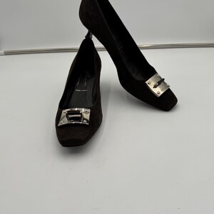 May include: A pair of black suede pumps with a square toe and a silver-tone metal buckle detail. The heels are approximately 3 inches high.