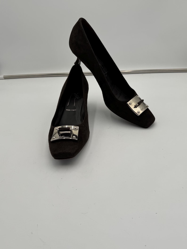 May include: A pair of black suede pumps with a square toe and a silver-tone metal buckle detail. The heels are approximately 3 inches high.