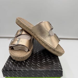 May include: Gold leather sandals with two adjustable buckles, resting on a box. The sandals have a woven jute sole and a metallic gold finish. The box is black with a green label that reads "Becca Gold Leather".
