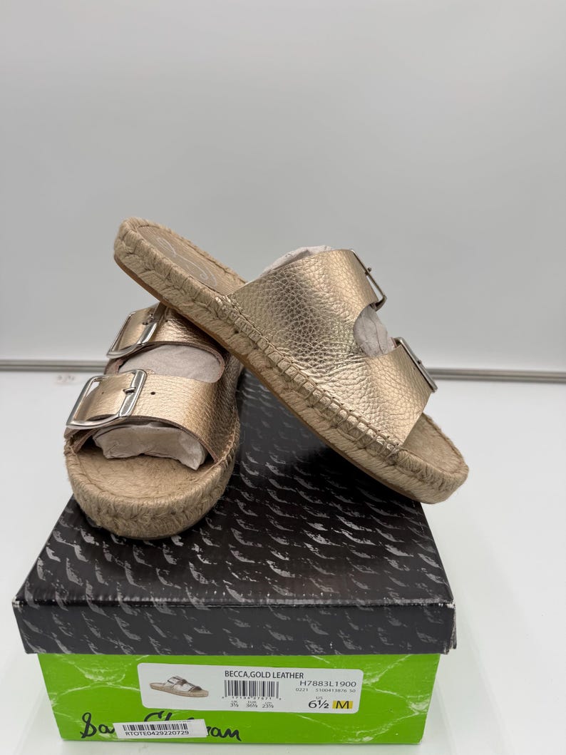 May include: Gold leather sandals with two adjustable buckles, resting on a box. The sandals have a woven jute sole and a metallic gold finish. The box is black with a green label that reads "Becca Gold Leather".
