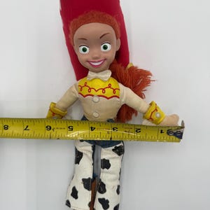 May include: A Jessie doll from the Toy Story franchise. The doll has red hair, a red hat, and a yellow shirt with red accents. She wears white pants with black cow spots and brown boots. The doll is approximately 7 inches wide.
