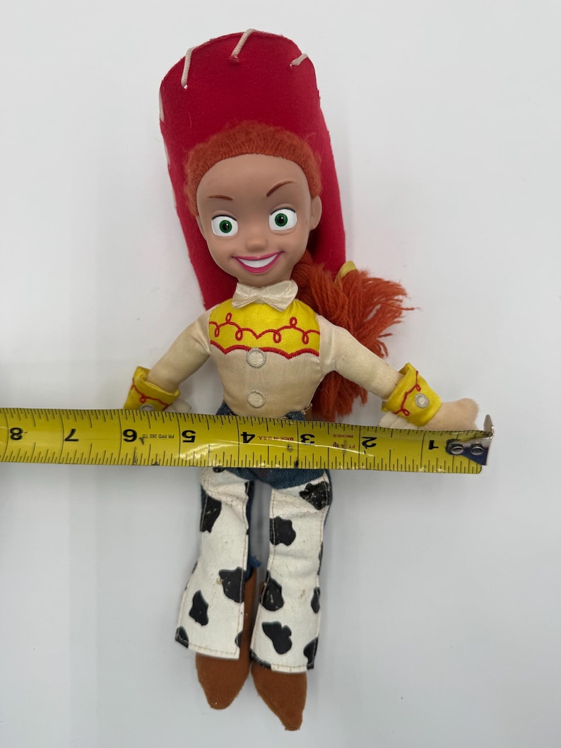 May include: A Jessie doll from the Toy Story franchise. The doll has red hair, a red hat, and a yellow shirt with red accents. She wears white pants with black cow spots and brown boots. The doll is approximately 7 inches wide.