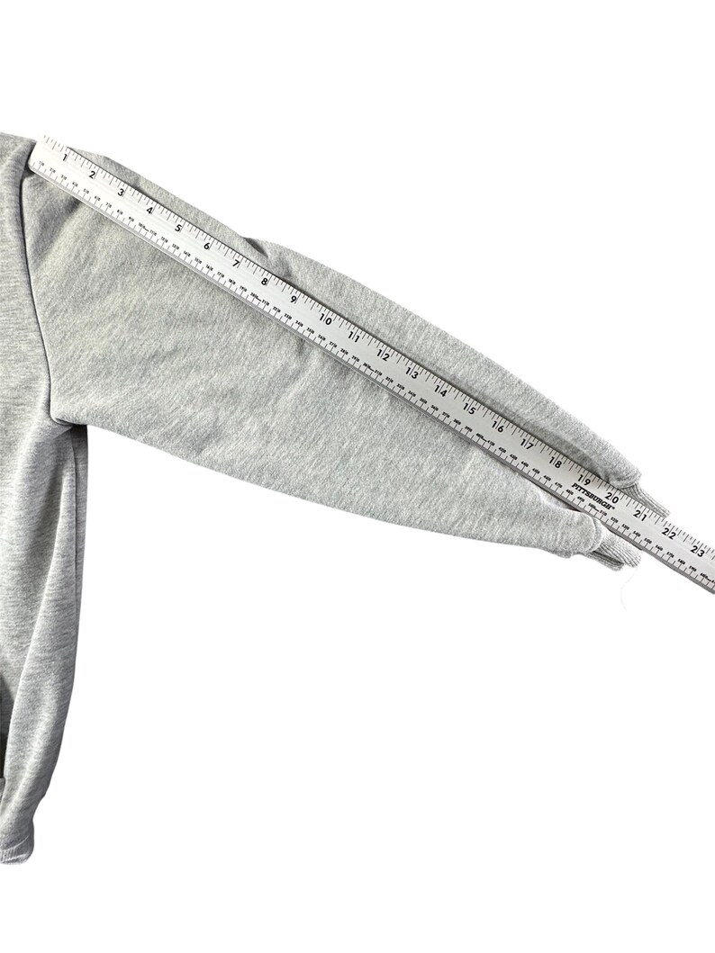 Puede incluir: Una manga de sudadera gris claro se mide con una regla. La manga est&aacute; hecha de una tela suave y texturizada. La regla muestra una medida de aproximadamente 56 cm desde la costura del hombro hasta el pu&ntilde;o.