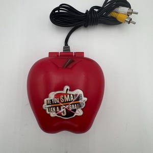 May include: A red plastic game console shaped like an apple, with a black cable and three AV connectors. The console has the text "ARE YOU SMARTER THAN A 5TH GRADER?" on a sticker.