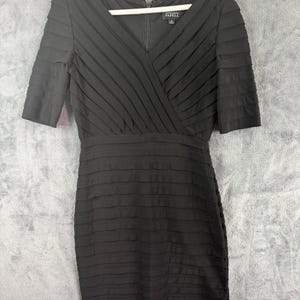 K&ouml;nnte beinhalten: Schwarzes knielanges Kleid mit V-Ausschnitt und kurzen &Auml;rmeln. Das Oberteil hat &uuml;berlappende Falten, der Rock horizontale Falten. Das Kleid h&auml;ngt an einem wei&szlig;en Kleiderb&uuml;gel vor grauem Hintergrund. Die Marke "Adrianna Papell" ist sichtbar.