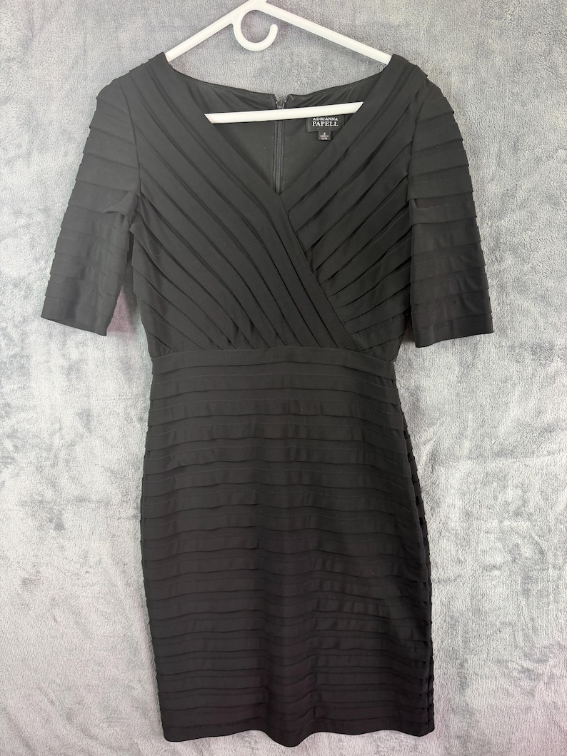 K&ouml;nnte beinhalten: Schwarzes knielanges Kleid mit V-Ausschnitt und kurzen &Auml;rmeln. Das Oberteil hat &uuml;berlappende Falten, der Rock horizontale Falten. Das Kleid h&auml;ngt an einem wei&szlig;en Kleiderb&uuml;gel vor grauem Hintergrund. Die Marke "Adrianna Papell" ist sichtbar.