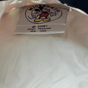 May include: A white fabric pouch with a black zipper featuring an interior label that reads Mickey & Co. Disney Honey Fashions New York, accompanied by a circular graphic of Mickey Mouse in red shorts and yellow shoes on a white background.