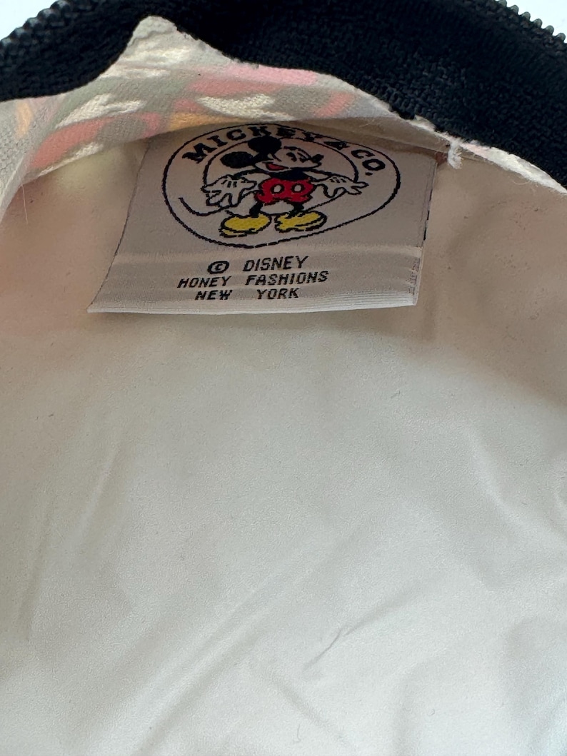 May include: A white fabric pouch with a black zipper featuring an interior label that reads Mickey & Co. Disney Honey Fashions New York, accompanied by a circular graphic of Mickey Mouse in red shorts and yellow shoes on a white background.