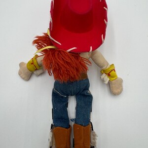 May include: A plush doll of a cowgirl character, featuring a red hat with white stitching, orange hair, and denim pants. The doll has brown boots, yellow wrist cuffs, and a tan body. A children's toy.