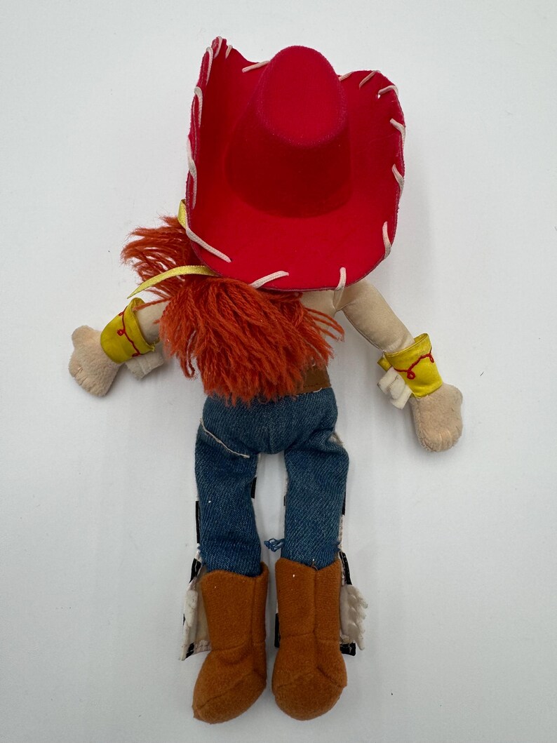 May include: A plush doll of a cowgirl character, featuring a red hat with white stitching, orange hair, and denim pants. The doll has brown boots, yellow wrist cuffs, and a tan body. A children's toy.