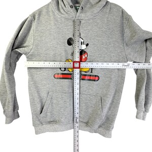 Puede incluir: Sudadera con capucha gris con un gr&aacute;fico de Mickey Mouse. El dise&ntilde;o presenta la cara y el cuerpo de Mickey, con un dise&ntilde;o rojo, negro y dorado en el pecho. La sudadera tiene un bolsillo delantero y mide aproximadamente 61 cm de largo.