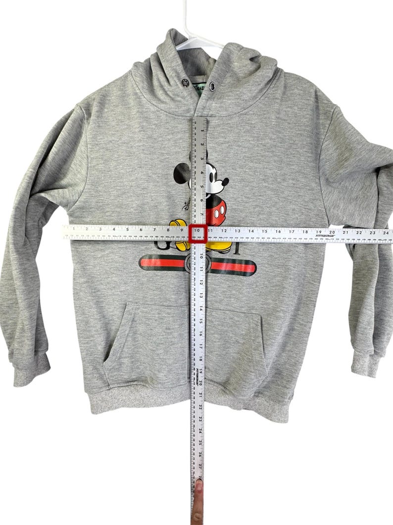 Puede incluir: Sudadera con capucha gris con un gr&aacute;fico de Mickey Mouse. El dise&ntilde;o presenta la cara y el cuerpo de Mickey, con un dise&ntilde;o rojo, negro y dorado en el pecho. La sudadera tiene un bolsillo delantero y mide aproximadamente 61 cm de largo.