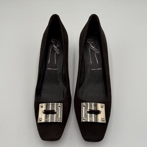 May include: A pair of dark brown suede flats with square toes. Each shoe features a silver-toned rectangular buckle on the vamp. The interior of the shoes is black, with text that reads "DONALD J. PLINER" and "Made in Spain".
