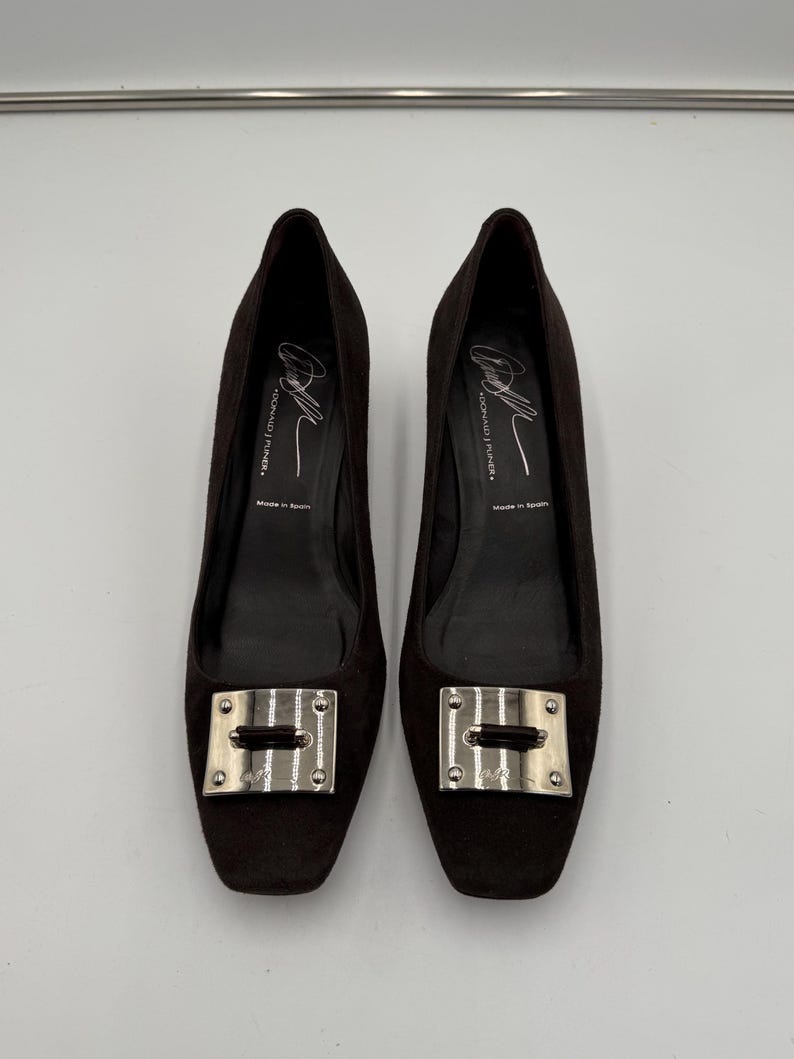 May include: A pair of dark brown suede flats with square toes. Each shoe features a silver-toned rectangular buckle on the vamp. The interior of the shoes is black, with text that reads "DONALD J. PLINER" and "Made in Spain".