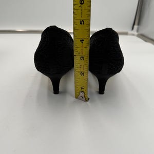 K&ouml;nnte beinhalten: Ein Paar schwarze Schuhe mit niedrigem Absatz und strukturiertem Blumenmuster. Die Abs&auml;tze sind etwa 5 cm hoch. Ein gelbes Lineal ist zwischen den Schuhen platziert und zeigt die H&ouml;he.