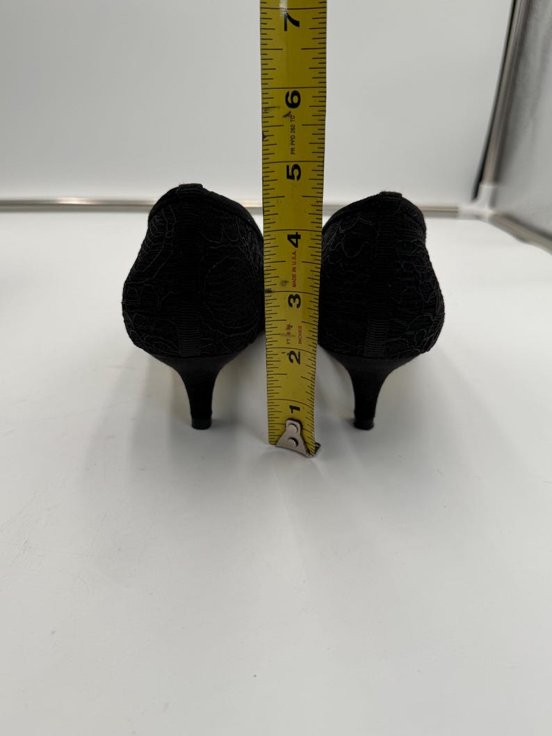 K&ouml;nnte beinhalten: Ein Paar schwarze Schuhe mit niedrigem Absatz und strukturiertem Blumenmuster. Die Abs&auml;tze sind etwa 5 cm hoch. Ein gelbes Lineal ist zwischen den Schuhen platziert und zeigt die H&ouml;he.