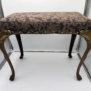 May include: An antique upholstered bench with a dark brown floral pattern on the seat. The legs are black with gold-colored ornate accents. The seat is rectangular and has brass-colored tacks along the bottom edge.