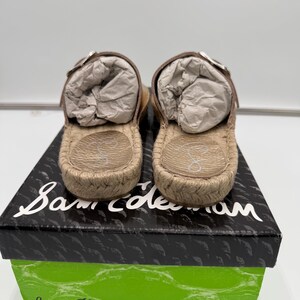 May include: A pair of tan espadrille sandals with brown leather straps and silver buckles, resting on a black and green box. The sandals have a woven jute sole and are filled with white paper.