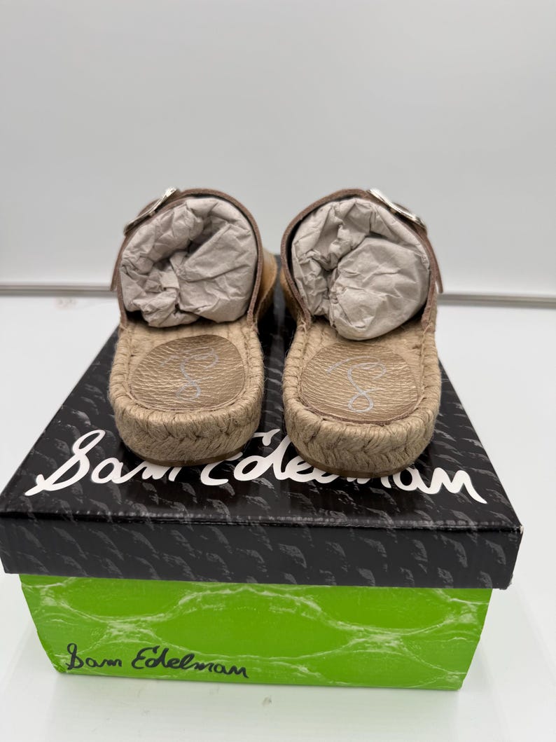 May include: A pair of tan espadrille sandals with brown leather straps and silver buckles, resting on a black and green box. The sandals have a woven jute sole and are filled with white paper.
