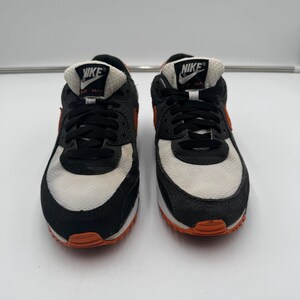 May include: Pair of Nike Air Max 90 sneakers. The shoes feature a black, white, and orange color scheme. The Nike logo is visible on the tongue. The sneakers have a classic design with a visible Air Max unit.