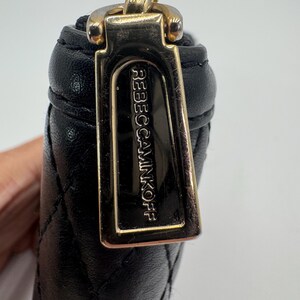Puede incluir: Primer plano de una cartera de cuero negro acolchado con un tirador dorado. El tirador de la cremallera es rectangular y presenta el nombre de la marca "REBECCAMINKOFF" en relieve. La cartera es parcialmente visible.