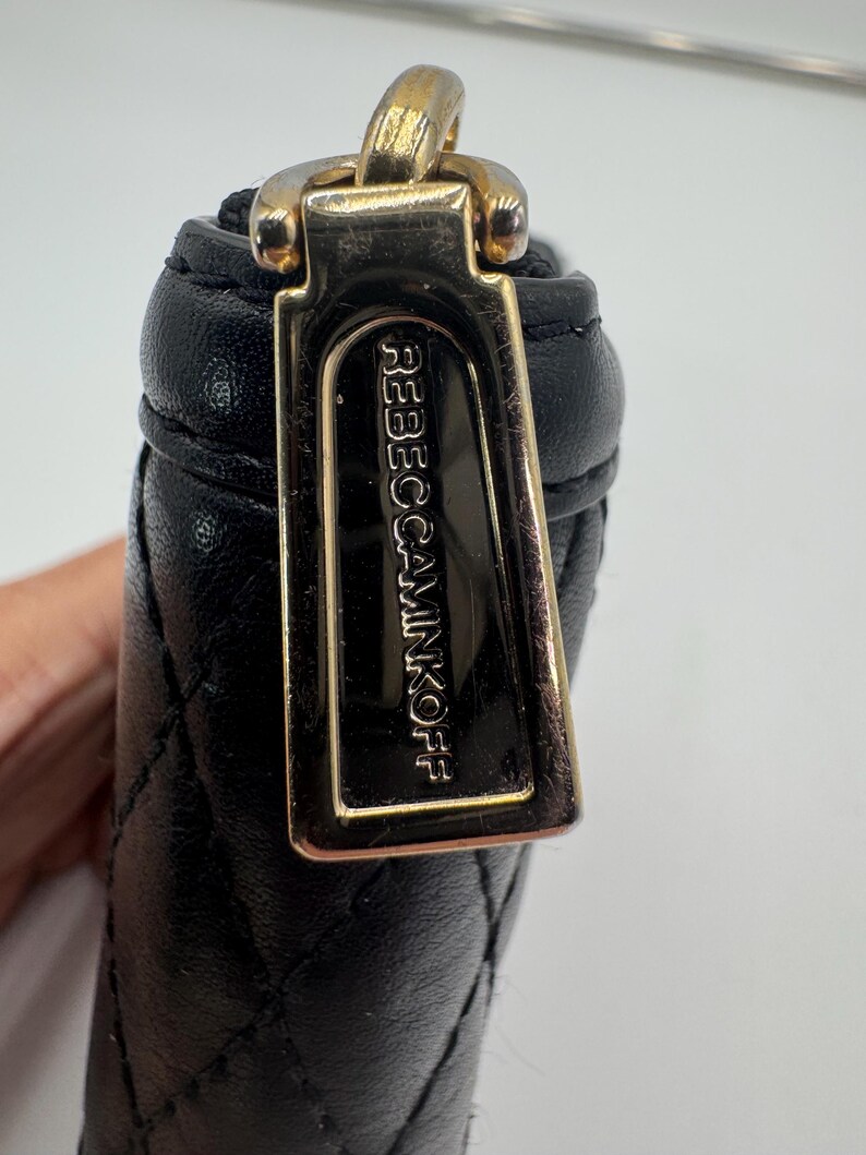 Puede incluir: Primer plano de una cartera de cuero negro acolchado con un tirador dorado. El tirador de la cremallera es rectangular y presenta el nombre de la marca "REBECCAMINKOFF" en relieve. La cartera es parcialmente visible.