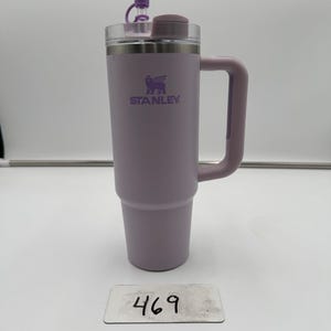 Op de afbeelding: Een lavendelkleurige Stanley ge&iuml;soleerde reismok met een handvat en een doorzichtig deksel. De mok heeft het Stanley-logo en een paarse stopper. Het nummer "469" staat op een klein wit label voor de mok.