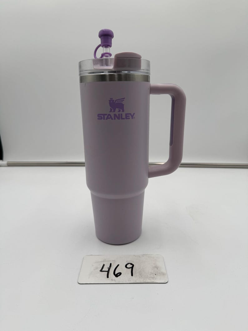 Op de afbeelding: Een lavendelkleurige Stanley ge&iuml;soleerde reismok met een handvat en een doorzichtig deksel. De mok heeft het Stanley-logo en een paarse stopper. Het nummer "469" staat op een klein wit label voor de mok.