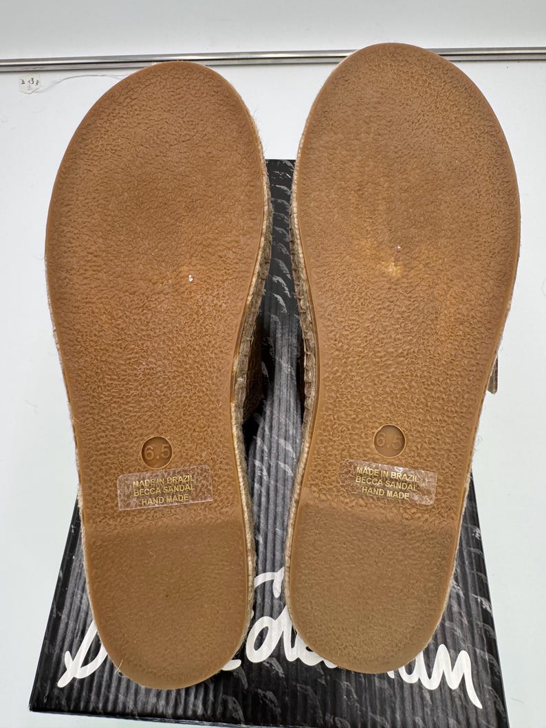 May include: Pair of espadrille sandals with a brown rubber sole, size 6.5. The sandals are handmade in Brazil. The soles are resting on a black and white patterned box.