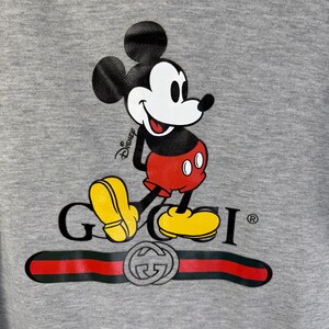 Puede incluir: Sudadera con capucha gris con un gr&aacute;fico de Mickey Mouse y el logotipo de Gucci. Mickey Mouse est&aacute; representado en su pose cl&aacute;sica, con pantalones cortos rojos y zapatos amarillos. El logotipo de Gucci es negro con una raya roja y verde.