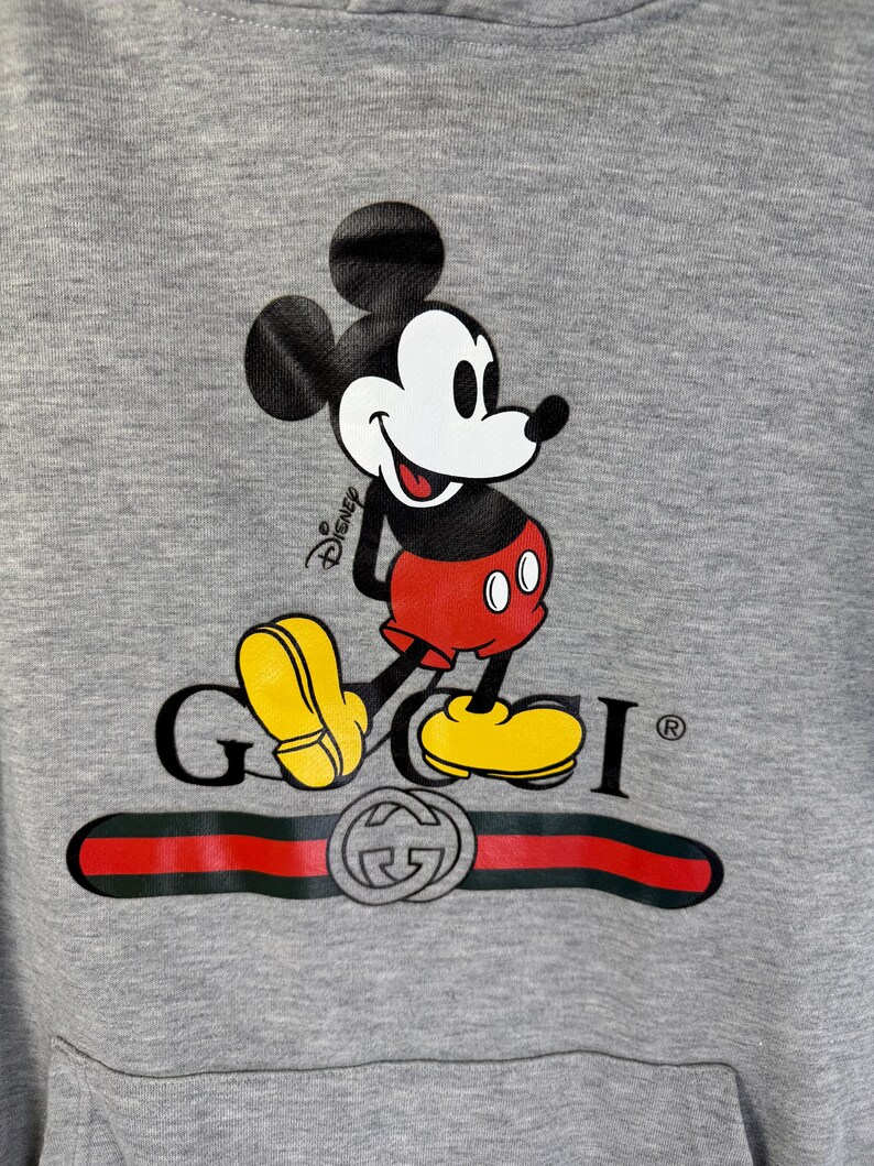 Puede incluir: Sudadera con capucha gris con un gr&aacute;fico de Mickey Mouse y el logotipo de Gucci. Mickey Mouse est&aacute; representado en su pose cl&aacute;sica, con pantalones cortos rojos y zapatos amarillos. El logotipo de Gucci es negro con una raya roja y verde.