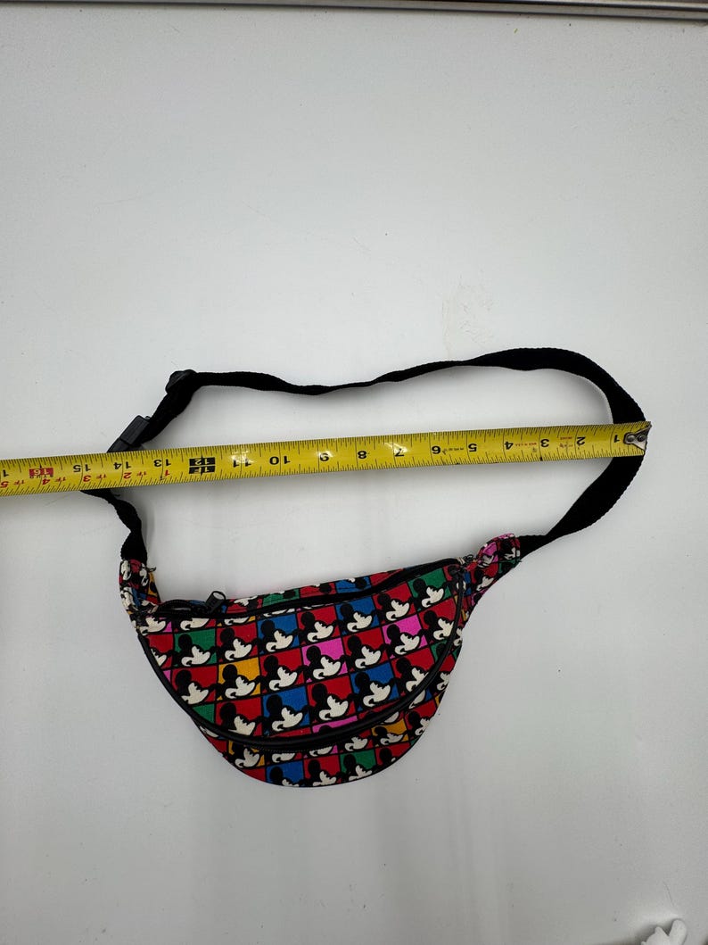 May include: A fanny pack featuring a repeating grid pattern of Mickey Mouse silhouettes in red, blue, yellow, and green squares. A black adjustable strap is attached. A yellow measuring tape shows the bag width is approximately 15 inches.