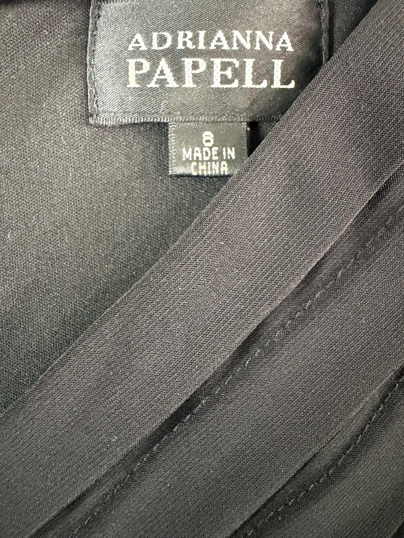 K&ouml;nnte beinhalten: Nahaufnahme eines schwarzen Kleidungsst&uuml;cks mit dem Etikett "ADRIANNA PAPELL". Das Etikett zeigt die Gr&ouml;&szlig;e 8 und "MADE IN CHINA". Der Stoff hat eine subtile Textur und diagonale Linien.