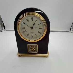 Puede incluir: Un reloj de cuarzo marrón oscuro con bisel y base dorados. La esfera tiene números romanos y la palabra "QUARTZ". El reloj tiene las iniciales "IB" en un cuadrado dorado. Es un artículo decorativo.