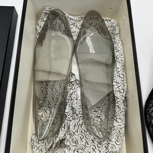 K&ouml;nnte beinhalten: Ein Paar transparente Schuhe mit spitzer Zehenpartie in einer schwarzen Box. Die Schuhe bestehen aus transparentem Material und sind mit wei&szlig;em Seidenpapier gef&uuml;llt. Die Box ist mit gemustertem Papier mit sich wiederholendem Design ausgekleidet.