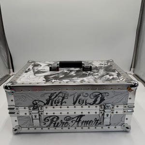 May include: Silver-toned metal case with a black handle and decorative rivets. The case features engraved artwork and the words "Kat Von D" and "Puro Amor" in script. The top of the case has a black and white illustration.