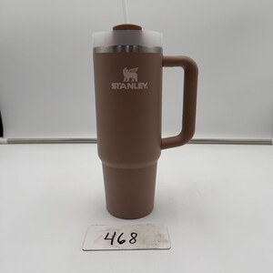Pu&ograve; includere: Un bicchiere Stanley marrone con manico e cannuccia bianca. Il bicchiere ha un coperchio trasparente e il logo Stanley &egrave; visibile. Il numero "468" &egrave; su una piccola etichetta bianca.