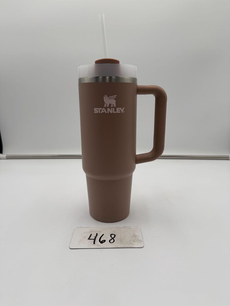 Pu&ograve; includere: Un bicchiere Stanley marrone con manico e cannuccia bianca. Il bicchiere ha un coperchio trasparente e il logo Stanley &egrave; visibile. Il numero "468" &egrave; su una piccola etichetta bianca.
