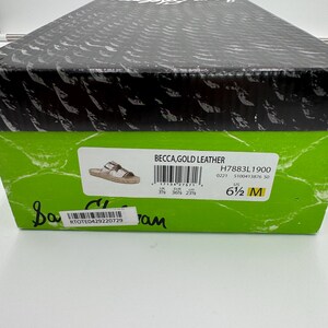 May include: A shoe box with a black lid and a green label. The label reads "BECCA, GOLD LEATHER" and includes the size "6 1/2 M". The box also has a barcode and the text "RTOTE0429220729".