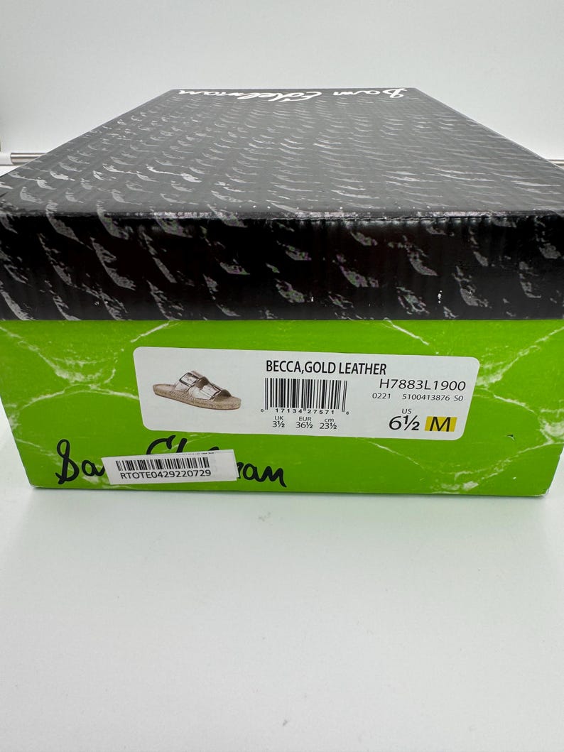 May include: A shoe box with a black lid and a green label. The label reads "BECCA, GOLD LEATHER" and includes the size "6 1/2 M". The box also has a barcode and the text "RTOTE0429220729".