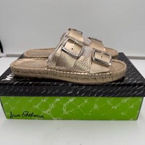 May include: Gold-colored sandals with two adjustable buckles. The sandals have a textured appearance and are set on a woven sole. The sandals are displayed on a black and green box with the brand name "Sam Edelman".