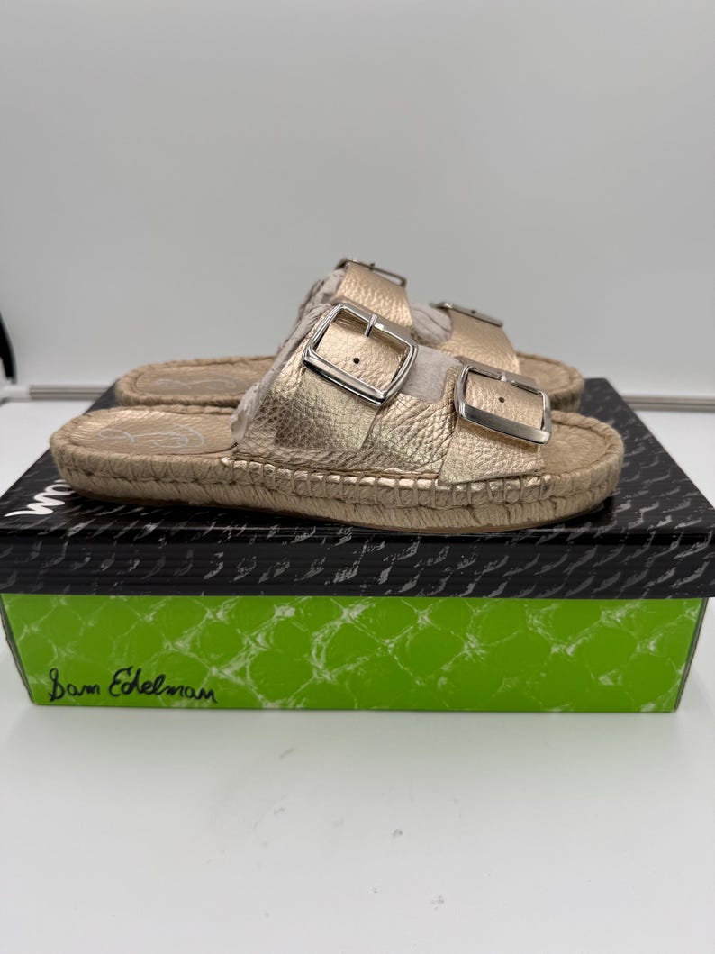 May include: Gold-colored sandals with two adjustable buckles. The sandals have a textured appearance and are set on a woven sole. The sandals are displayed on a black and green box with the brand name "Sam Edelman".