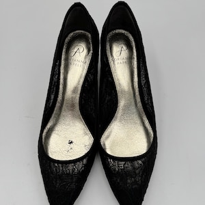 K&ouml;nnte beinhalten: Ein Paar schwarze Pumps mit Spitze und spitzer Zehenpartie. Die Schuhe haben eine silberfarbene Innensohle und der Markenname "Adrianna Papell" ist innen sichtbar. Die Abs&auml;tze sind niedrig und das Design ist elegant.