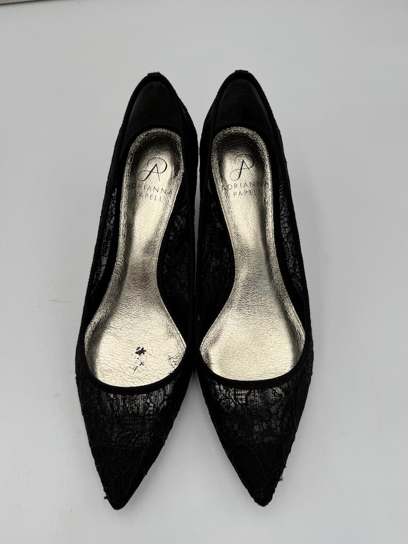 K&ouml;nnte beinhalten: Ein Paar schwarze Pumps mit Spitze und spitzer Zehenpartie. Die Schuhe haben eine silberfarbene Innensohle und der Markenname "Adrianna Papell" ist innen sichtbar. Die Abs&auml;tze sind niedrig und das Design ist elegant.