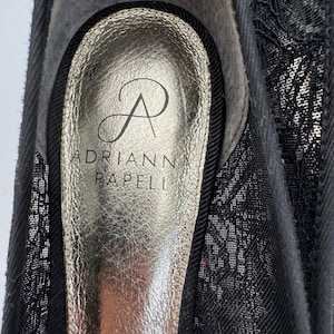 K&ouml;nnte beinhalten: Nahaufnahme eines schwarzen Schuhs mit goldener Innensohle. Die Innensohle tr&auml;gt den Markennamen "ADRIANNA PAPELL" und ein stilisiertes "A"-Logo. Der Schuh hat einen schwarzen Spitzen-Overlay.