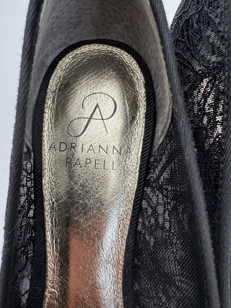 K&ouml;nnte beinhalten: Nahaufnahme eines schwarzen Schuhs mit goldener Innensohle. Die Innensohle tr&auml;gt den Markennamen "ADRIANNA PAPELL" und ein stilisiertes "A"-Logo. Der Schuh hat einen schwarzen Spitzen-Overlay.