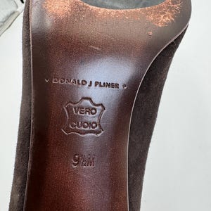 May include: Close-up of a brown suede high heel shoe sole. The sole is marked with "DONALD J PLINER", "VERO CUOIO", and size 9 1/2 M. The heel and sole show signs of wear.