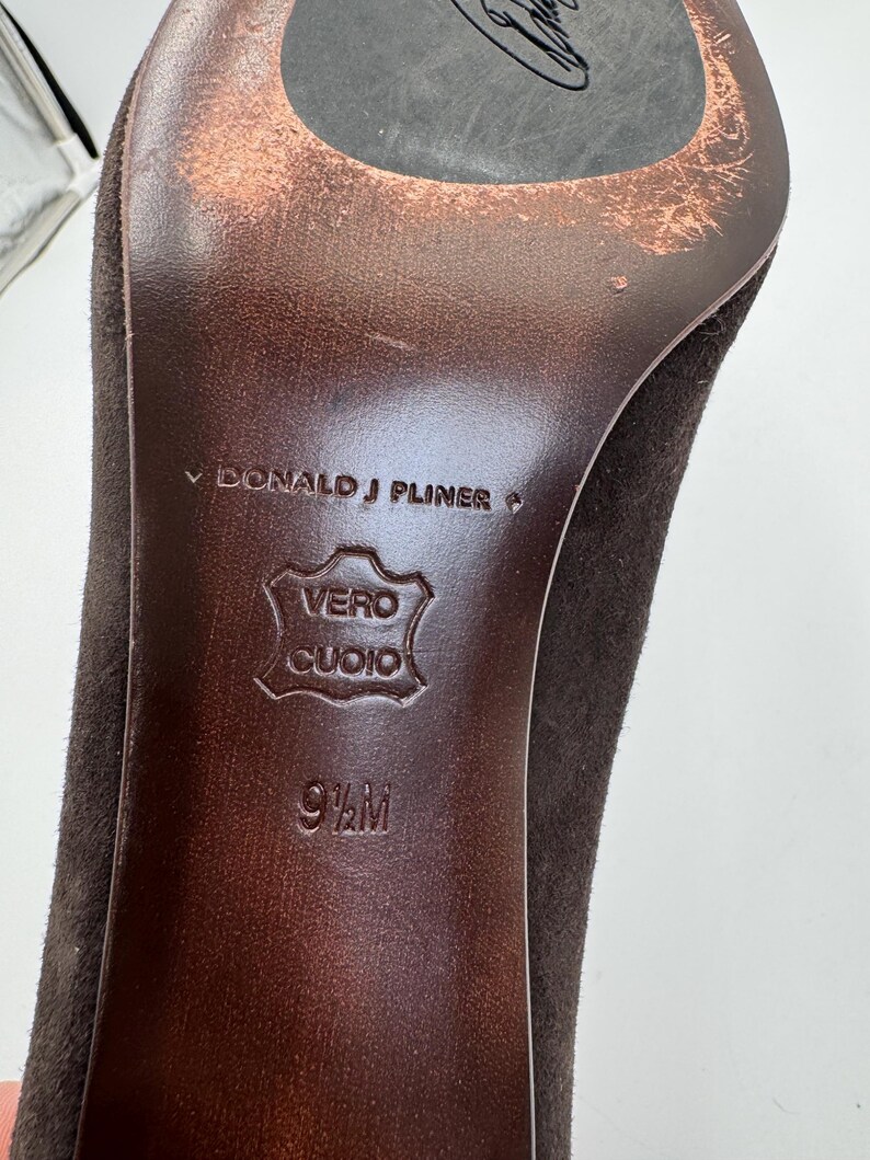 May include: Close-up of a brown suede high heel shoe sole. The sole is marked with "DONALD J PLINER", "VERO CUOIO", and size 9 1/2 M. The heel and sole show signs of wear.