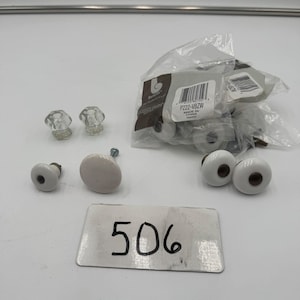 May include: A collection of cabinet knobs including two clear faceted glass knobs and several white ceramic knobs with dark centers. Some knobs are loose, while others are contained in a clear plastic bag with a label reading P222-VBZW. A white rectangular card with the handwritten number 506 is placed in the foreground.